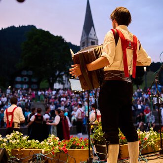 © Kleinwalsertal Tourismus | Oliver Farys Ein Musiker spielt auf einem Akkordeon vor einer großen Menschenmenge. Die Atmosphäre ist festlich, umgeben von einer schönen Naturlandschaft. | © Kleinwalsertal Tourismus | Oliver Farys