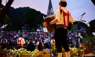 Ein Musiker spielt auf einem Akkordeon vor einer großen Menschenmenge. Die Atmosphäre ist festlich, umgeben von einer schönen Naturlandschaft. | © Kleinwalsertal Tourismus | Oliver Farys