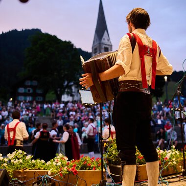 Ein Musiker spielt auf einem Akkordeon vor einer großen Menschenmenge. Die Atmosphäre ist festlich, umgeben von einer schönen Naturlandschaft. | © Kleinwalsertal Tourismus | Oliver Farys