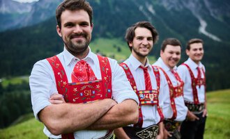 Vier Männer in traditioneller Tracht stehen vor einer malerischen Berglandschaft. Sie lächeln freundlich in die Kamera, die Umgebung ist grün und hügelig. | © Kleinwalsertal Tourismus | Oliver Farys
