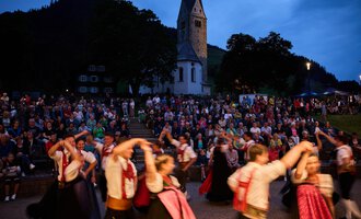 Eine festliche Tanzaufführung findet vor einer Kirche statt. Im Hintergrund ist eine große Menschenmenge zu sehen, die zuschaut. | © Kleinwalsertal Tourismus | Oliver Farys
