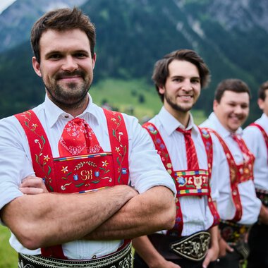 Vier Männer in traditioneller Tracht stehen vor einer malerischen Berglandschaft. Sie lächeln freundlich in die Kamera. | © Kleinwalsertal Tourismus | Oliver Farys