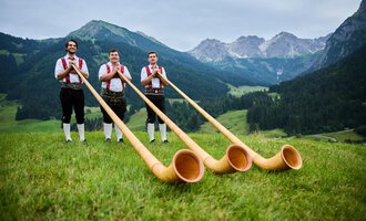 Three men in traditional attire stand in a green meadow with an impressive mountain landscape in the background. They hold large alpine horns in their hands. | © Kleinwalsertal Tourismus eGen | Oliver Farys