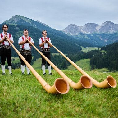 Three men in traditional attire stand in a green meadow with an impressive mountain landscape in the background. They hold large alpine horns in their hands. | © Kleinwalsertal Tourismus eGen | Oliver Farys