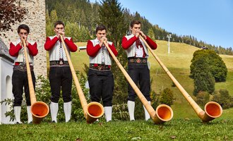 Four young men in traditional clothing are playing alpine horns in a picturesque landscape. In the background, green meadows and trees can be seen. | © Kleinwalsertal Tourismus | Frank Drechsel