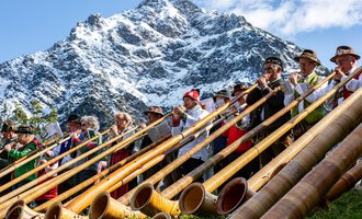 A group of musicians is playing alphorn in the mountains. In the background, snow-covered peaks and a blue sky can be seen. | © Kleinwalsertal Tourismus eGen | Frank Drechsel