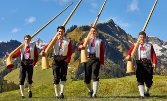 Four men in traditional costumes are carrying large alpine horns. In the background, green meadows and impressive mountains can be seen. | © Kleinwalsertal Tourismus | Frank Drechsel