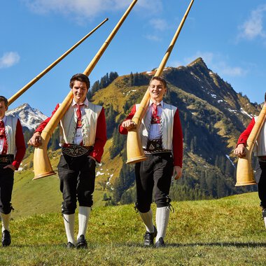Four men in traditional costumes are carrying large alpine horns. In the background, green meadows and impressive mountains can be seen. | © Kleinwalsertal Tourismus | Frank Drechsel