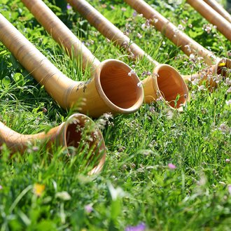 A series of alphorns lies in the grass. The horns are made of wood and surrounded by colorful flowers. | © OBERSTDORF · KLEINWALSERTAL BERGBAHNEN