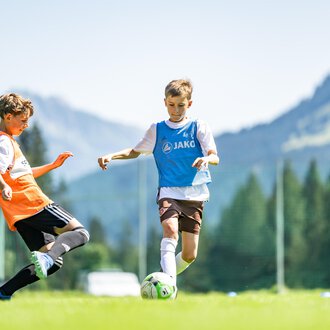 © Fußballschule Anstoß Junge Fußballspieler auf einem grünen Spielfeld mit Bergen im Hintergrund. Ein Spieler dribbelt den Ball, während ein anderer ihn versucht zu stoppen. | © Fußballschule Anstoß