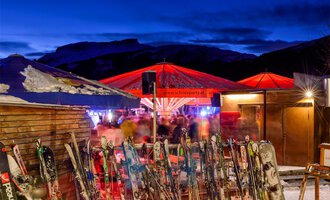 A ski lodge with red sun umbrellas in a wintry setting. In the foreground, several skis are visible, and in the background, people can be seen enjoying the atmosphere. | © Plattform Kleinwalsertal | Michael Monschau