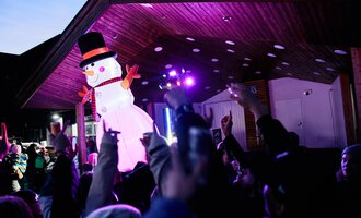 A festive event with a large snowman puppet and many celebrating people. Colorful lights illuminate the scene. | © Kleinwalsertal Tourismus | Oliver Farys