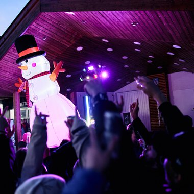 A festive event with a large snowman puppet and many celebrating people. Colorful lights illuminate the scene. | © Kleinwalsertal Tourismus | Oliver Farys