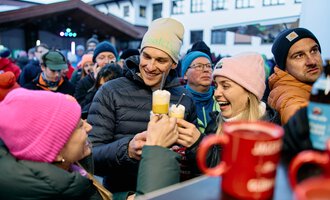 A cheerful group of people toasts with drinks. They are wearing warm jackets and hats and are standing in a lively crowd. | © Kleinwalsertal Tourismus | Oliver Farys