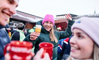 A cheerful group of people in winter clothing is celebrating outdoors. A woman with a pink hat is holding a drink and smiling. | © Kleinwalsertal Tourismus | Oliver Farys