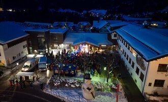 A festive event in a snowy village at night. People are gathered around a stage while a large snowman stands nearby. | © Kleinwalsertal Tourismus eGen | Oliver Farys