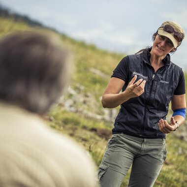 Eine Frau spricht mit einem Mann im Freien. Sie hält einen Gegenstand in der Hand und lächelt freundlich. | © Kleinwalsertal Tourismus | Oliver Farys