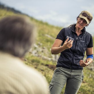 A woman is talking to a man outdoors. She is holding an object in her hand and smiling kindly. | © Kleinwalsertal Tourismus | Oliver Farys