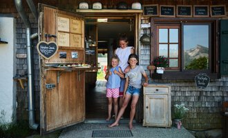 Eine einladende Eingangstür zu einem kleinen Laden, umgeben von Holz und Grün. Drei Kinder stehen fröhlich davor und lächeln in die Kamera. | © Kleinwalsertal Tourismus | Oliver Farys