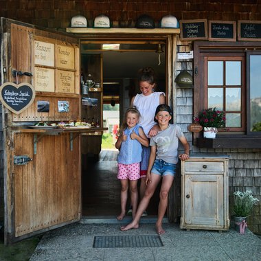 Eine einladende Eingangstür zu einem kleinen Laden, umgeben von Holz und Grün. Drei Kinder stehen fröhlich davor und lächeln in die Kamera. | © Kleinwalsertal Tourismus | Oliver Farys