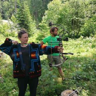 Zwei Personen im Wald beim Bogenschießen. Die Frau trägt einen bunten Pullover und zielt mit einem Bogen, während der Mann in einem grünen T-Shirt hinter ihr steht. | © Wildnisschule Kleinwalsertal | Ronny Katz