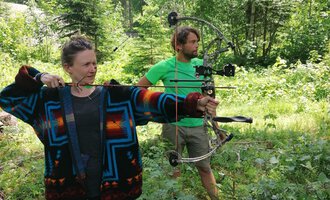 Zwei Personen im Wald beim Bogenschießen. Die Frau trägt einen bunten Pullover und zielt mit einem Bogen, während der Mann in einem grünen T-Shirt hinter ihr steht. | © Wildnisschule Kleinwalsertal | Ronny Katz