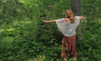Eine Frau steht im Wald und zielt mit einem Bogen. Um sie herum sind üppige grüne Pflanzen und Bäume.