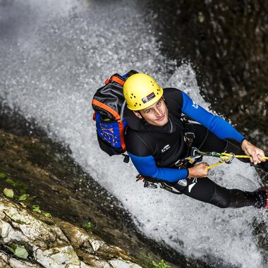 A man is climbing a steep rock wall in a waterfall. He is wearing a helmet and climbing gear. | © Bergschule Kleinwalsertal | Dominik Berchtold