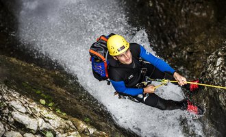 A man is climbing a steep rock wall in a waterfall. He is wearing a helmet and climbing gear. | © Bergschule Kleinwalsertal | Dominik Berchtold