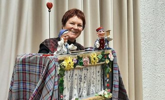 A woman presents a puppet show with two handcrafted puppets. The background is adorned with a bright curtain and colorful decorations. | © Marie Luise Kaiser | Veronika Senn