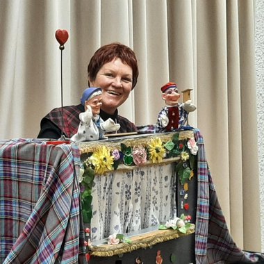 A woman presents a puppet show with two handcrafted puppets. The background is adorned with a bright curtain and colorful decorations. | © Marie Luise Kaiser | Veronika Senn