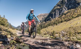 Two cyclists ride on a hiking trail through the mountains. The landscape is green and sunny under a clear blue sky. | © Kleinwalsertal Tourismus | Bastian Morell