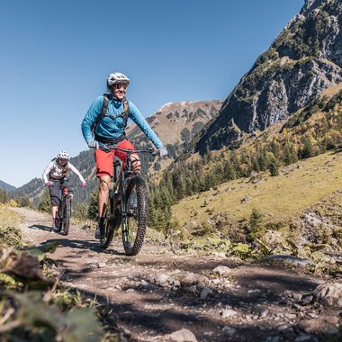 Two cyclists ride on a hiking trail through the mountains. The landscape is green and sunny under a clear blue sky. | © Kleinwalsertal Tourismus | Bastian Morell