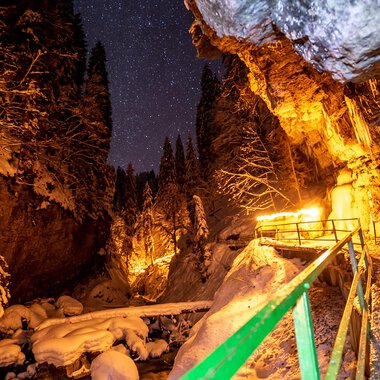 A narrow, snow-covered path winds through a deeply snow-laden gorge. The night sky is dotted with stars, while warm lights illuminate the surroundings. | © Breitachklammverein eG | Dominik Berchtold