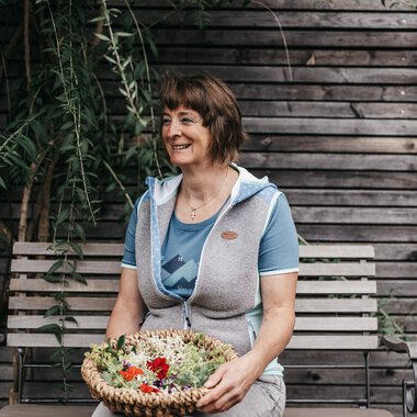 A smiling woman is sitting on a bench holding a basket of fresh plants. In the background, wooden slats and green plants can be seen. | © Naturhotel Chesa Valisa