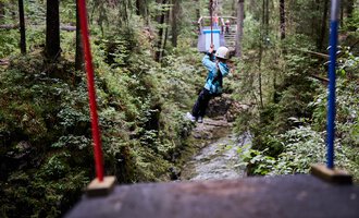 A child swings on a zip line between trees in a green forest. Surrounded by nature and a narrow stream below. | © Kleinwalsertal Tourismus | Oliver Farys