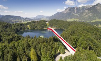An impressive viewpoint bridge over a clear lake, surrounded by green forests and mountains. The sky is blue with some clouds. | © Skiflugschanze Oberstdorf | Eren Karaman