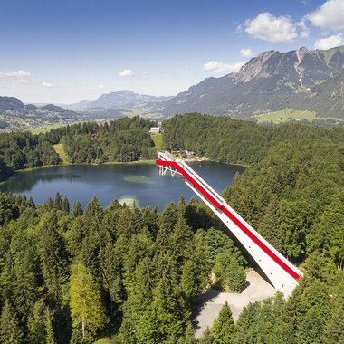 An impressive viewpoint bridge over a clear lake, surrounded by green forests and mountains. The sky is blue with some clouds. | © Skiflugschanze Oberstdorf | Eren Karaman