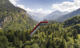 An impressive observation platform that extends above the trees, surrounded by majestic mountains and dense forests. The sky is clear and shows some clouds. | © Skiflugschanze Oberstdorf | Eren Karaman