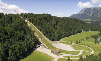 An impressive ski jump surrounded by green forests and mountains. The landscape is picturesque and showcases a mix of nature and sports facilities. | © Skiflugschanze Oberstdorf | Eren Karaman