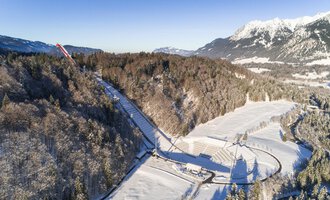 A snow-covered landscape with trees and a clear sky. In the background, mountains can be seen that complement the wintry scenery. | © Skiflugschanze Oberstdorf | Eren Karaman