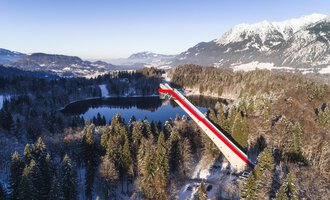 A long bridge with red and white stripes spans a tranquil, snow-covered valley and a lake. In the background, majestic mountains can be seen, glowing in the winter sun. | © Skiflugschanze Oberstdorf | Eren Karaman