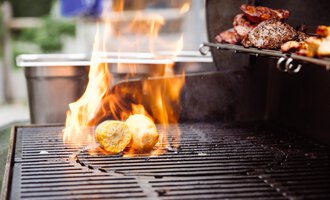 Ein Grill mit Flammen, auf dem Lebensmittel gekocht werden. Man sieht gegrillte Kartoffeln und Fleisch. | © Hotel Auenhütte | Eva Backes