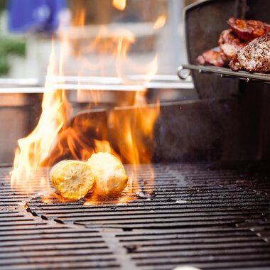 Ein Grill mit Flammen, auf dem Lebensmittel gekocht werden. Man sieht gegrillte Kartoffeln und Fleisch. | © Hotel Auenhütte | Eva Backes