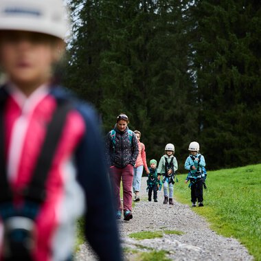 A group of children is hiking on a gravel path through a forest. They are wearing helmets and are well-equipped for an adventure in nature. | © Kleinwalsertal Tourismus | Oliver Farys