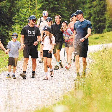Two hikers walk along a path in the mountains. The sun is shining and the landscape is green and golden. | © Kleinwalsertal Tourismus eGen - Fotograf: Oliver Farys