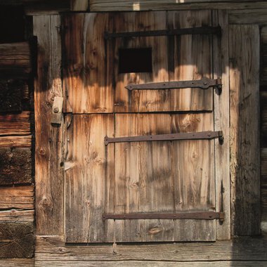Eine alte Holzlatttür mit Eisenbeschlägen. Die Tür hat ein kleines Fenster und ist von einer rustikalen Wand umgeben. | © R. Koeberle