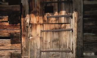 An old wooden plank door with iron fittings. The door has a small window and is surrounded by a rustic wall. | © R. Koeberle