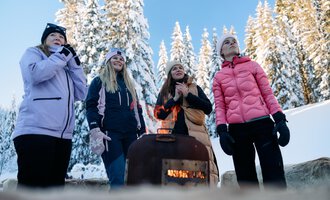 A group of women stands around a small heater in a snowy winter landscape. In the background, snow-covered trees and a clear blue sky can be seen. | © Kleinwalsertal Tourismus | Oliver Farys