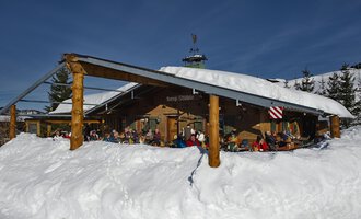 A cozy mountain cabin in the snow with many people on the terrace. The surroundings are wintry and the sun is shining in the blue sky. | © Bergstüble | Michaela Beck Dornach